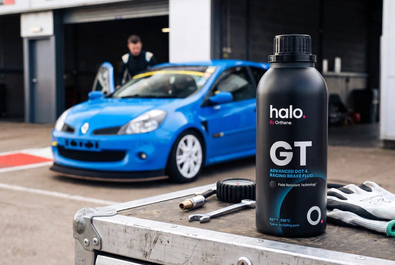 A bottle of halo GT racing grease fluid in the foreground on a metal toolbox, with a blue race car and a garage workshop in the background.